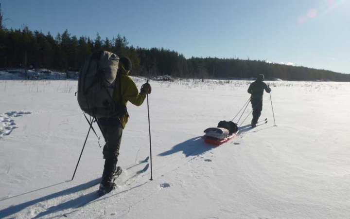 In the foreground, a person wearing a backpack uses trekking poles as they move over a vast snowy landscape. In front of them, another person on cross country skis pulls a small sled. 